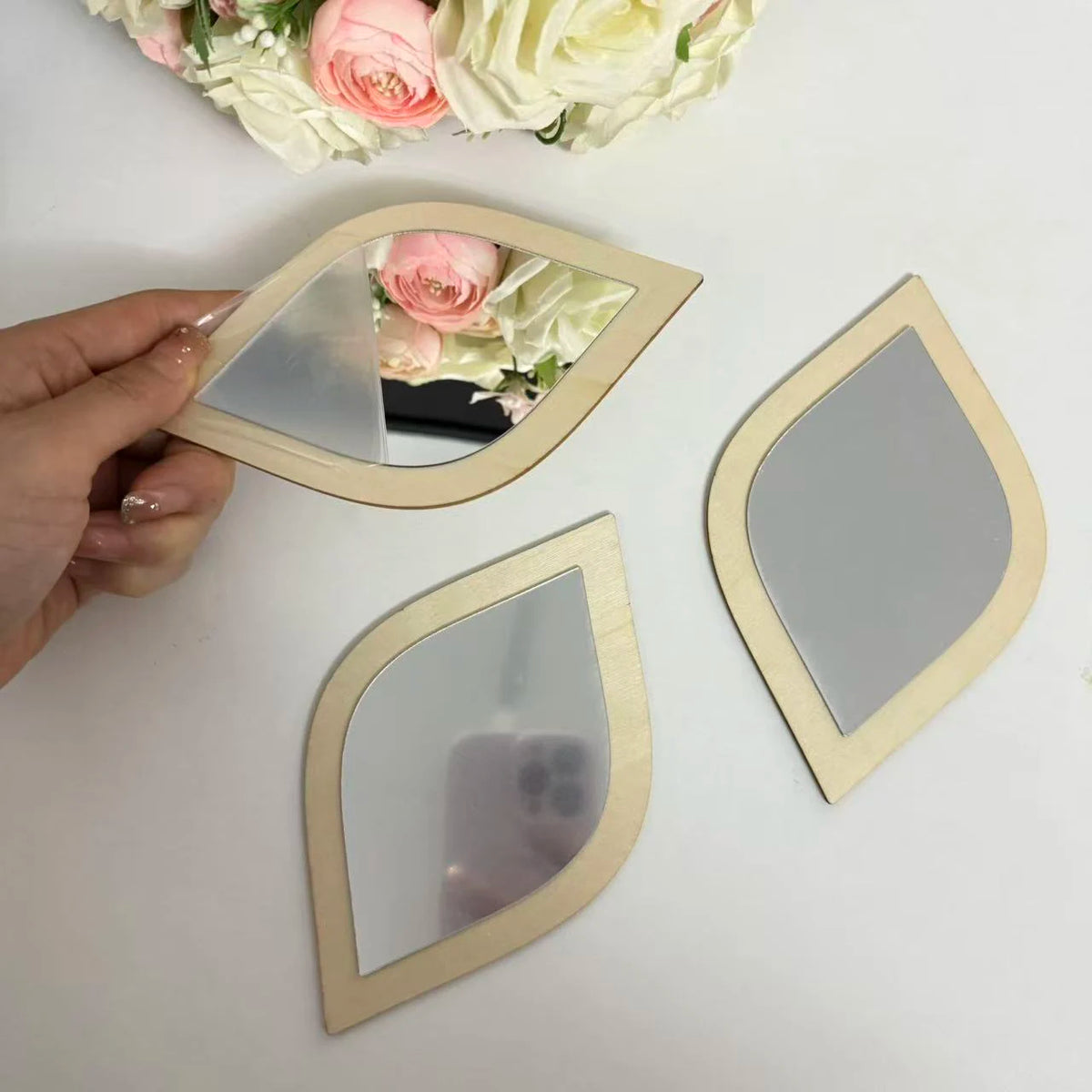 Hand holding a leaf-shaped wood frame mirror showing three sizes arranged on a white surface